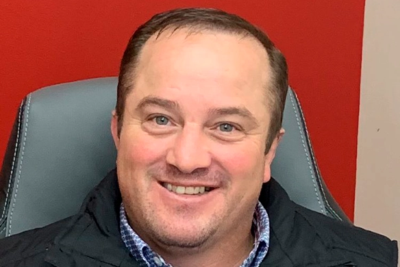 Portrait of Rick Stimpson smiling in front of a red wall and seated in an office chair