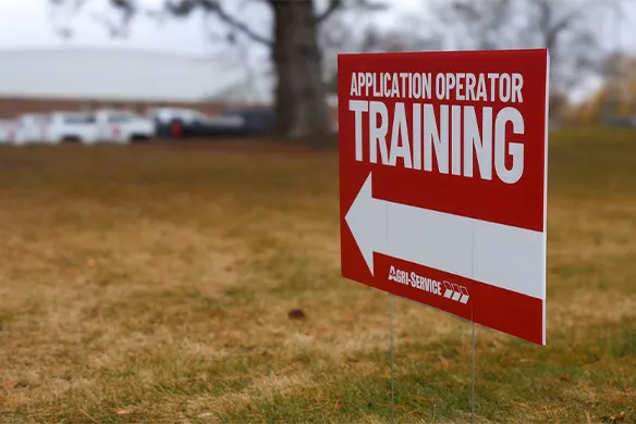 Agri-Service Application Operator training yard sign, with arrow pointing toward parked service trucks in the background.